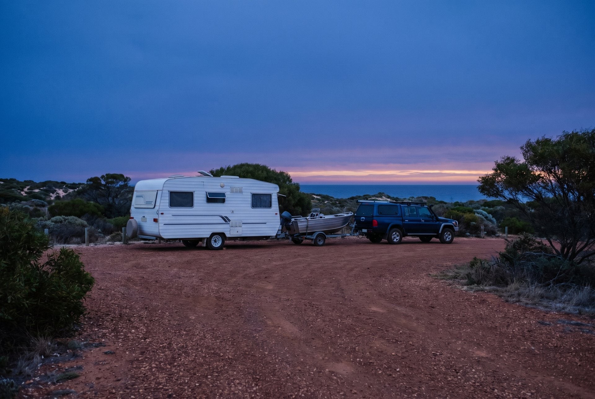 A caravan, a boat on its trailer and a 4WD parked at a coastal camp at dusk